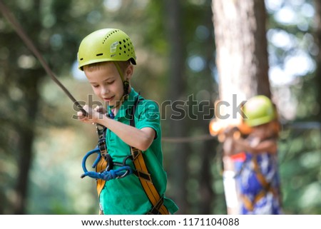 Young cute blond smiling child boy in safety harness and helmet attached with carbine to cable moves along rope way on bright sunny bokeh background. Sport, game, leisure, outdoor activity concept.