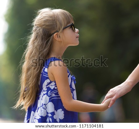 Back view of pretty young child girl with beautiful long blond hair in fashionable blue dress and sunglasses holding hand of young woman outdoors on blurred colorful sunny summer bokeh background.