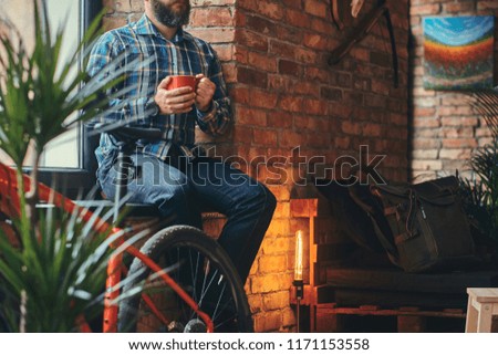 Handsome bearded hipster male in a blue fleece shirt and jeans holds a cup of morning coffee while sitting on a window sill at a studio with a loft interior.