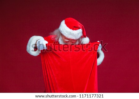 Christmas. Santa Claus in white gloves opened the bag with gifts and looks into it. Offers to take the gifts. Isolated on red background.