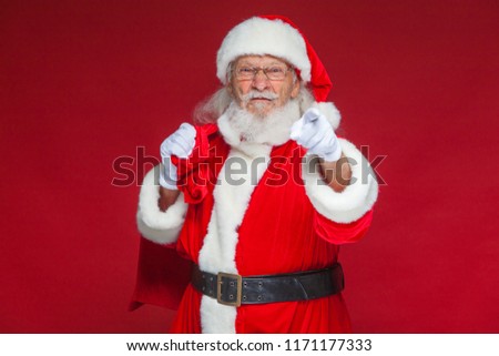 Christmas. Santa Claus in white gloves with a dissatisfied face expression with a bag of gifts behind his back points his index finger into the camera. Isolated on red background.
