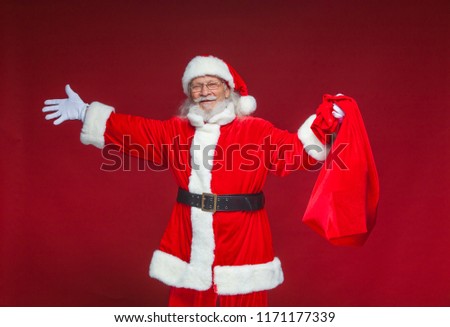 Christmas. Kind smiling Santa Claus spread his hands to the sides. In one hand he is holding a red bag with gifts. Isolated on red background.