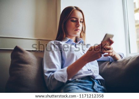 Thoughtful hipster girl in trendy outfit resting after work at comfortable sofa with mobile phone in hand, pondering woman booking hotel online via contemporary smartphone with 4g internet indoors