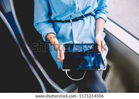 Cropped image of woman sitting inside bus using modern digital tablet and earphones watching video online, female listening audiobook via app on portable pc connected to free wifi in transport