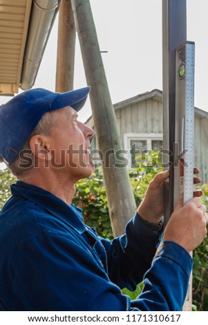 A middle-aged man builder in a blue jacket and blue cap places a black iron pillar on the level