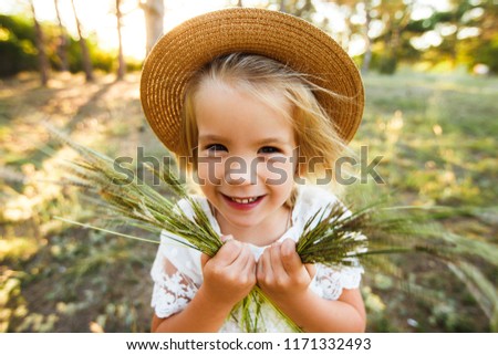 A cute baby girl in a straw hat and white dress is sitting on the grass.