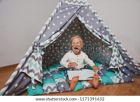 Happy boy smiling and holding book sitting in wigwam tent in the room