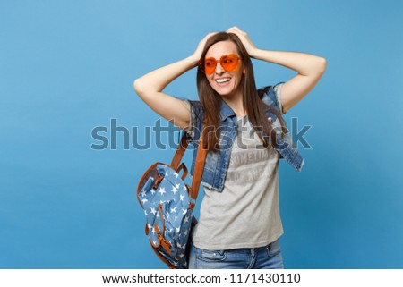 Young cheerful attractive woman student with backpack in orange heart glasses looking aside keeping hands on head isolated on blue background. Education in university. Copy space for advertisement