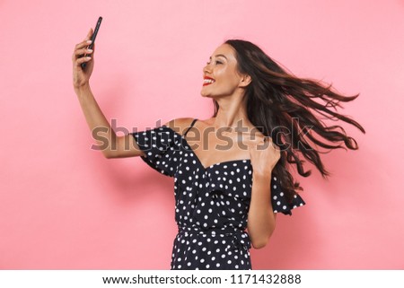 Image of Cheerful brunette woman in dress making selfie on smartphone over pink background