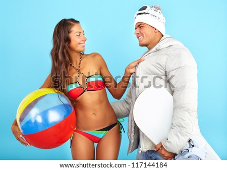 Portrait of happy girl in bikini with ball looking at handsome man in winterwear holding snowboard