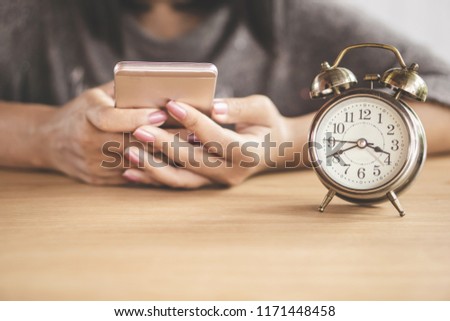 wasting time at work concept with woman using smart phone with alarm clock on desk