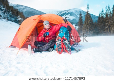 A man drinks from a mug near the tent in the winter. Tent, backpack, tracking sticks and snowshoes on the background of the mountains. Camping in the snow. Active rest and mountain climbing in winter.
