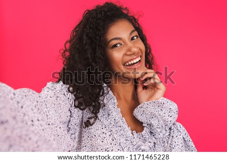 Portrait of a happy young african woman in summer dress standing isolated over pink background, taking a selfie with mobile phone
