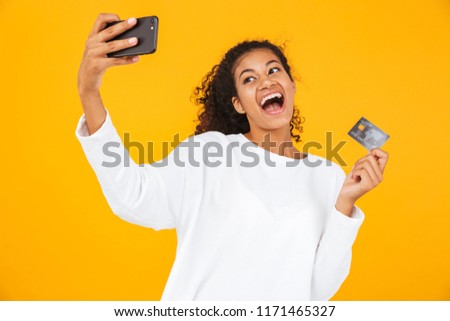 Portrait of a smiling young african woman standing isolated over yellow background, holding plastic credit card and taking selfie