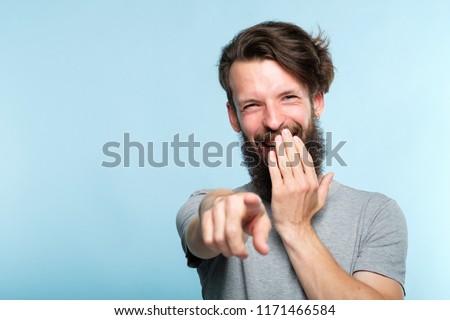 omg. ha-ha. bearded hipster man mocking and laughing at viewer pointing finger and covering mouth. sneer humiliation and psycological abuse concept. portrait of a grinning guy on blue background. Royalty-Free Stock Photo #1171466584