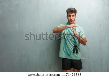 Young fitness man against a grunge wall tired and bored, making a timeout gesture, needs to stop because of work stress, time concept. Holding a jump rope.