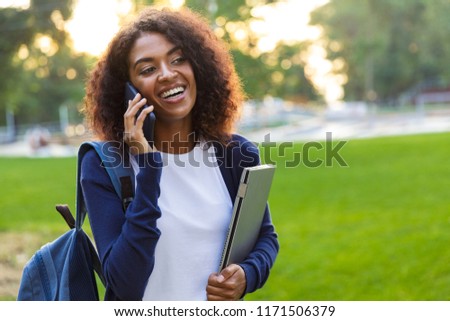 Image of young beautiful african woman student walking in the park holding laptop talking by mobile phone.