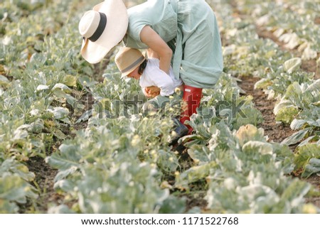 Mom in green linen dress and red rubber boots with son wearing white linen shirt and blue rubber boots are having fan in the cabbage field