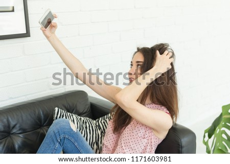 Technology, beauty and people concept - young smiling woman trying to make photo using smartphone and lying on sofa at home touching her hair.