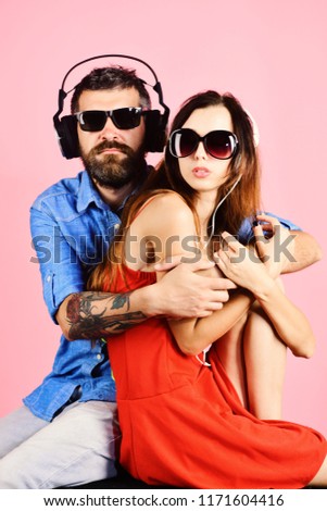 Music fans with calm faces enjoy music. Man with beard hugs pretty girl on pink background. Couple in love wears plastic headphones and sunglasses. Pleasure, music and creative lifestyle concept.