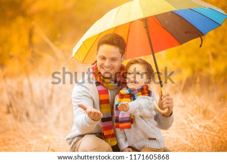 A little boy and his father are under a colorful umbrella. Family in warm clothes and colorful scarves. the kid catches the rain drops with his palm
