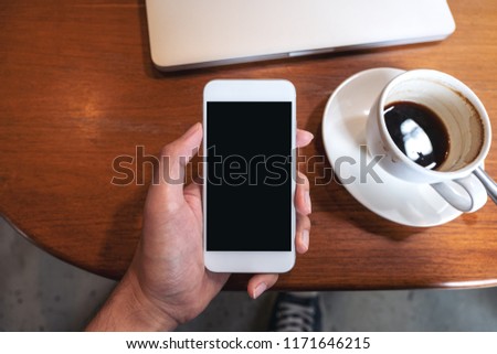 Top view mockup image of hand holding white mobile phone with blank black desktop screen with coffee cup and laptop on wooden table in cafe