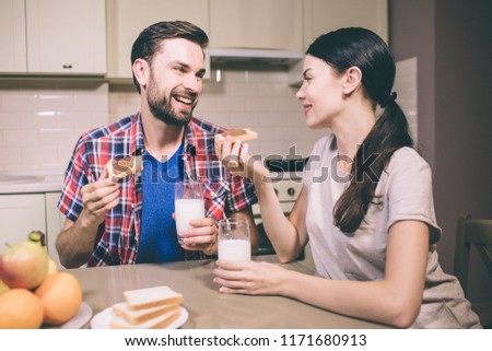 A picture of nice couple sits at table and look at each other. They hold toasts with chocolate paste on it. Also there are glasses of milk in their hands. People look happy.