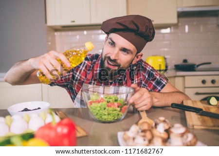 A picture of cook standing at table and holds bottle of sunflower oil. He looks at it and pouring some oil into bowl with salad. Man is doing that careful. Also he holds another hand on bowl.