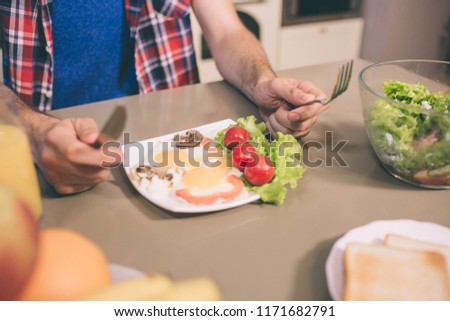 A picture of tasty omlet with letuce and cherry tomatoes on plate on table. Guy holds knife and fork in hands. He is ready to eat it.