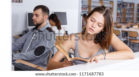 Attractive girl preparing for exam in college library, reading book and using pc for searching information