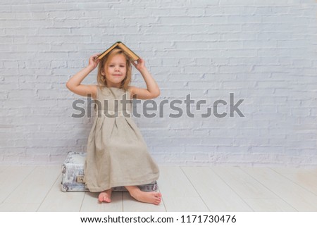 girl, baby in dress on white brick wall background with suitcase, with a book