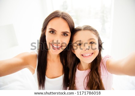 Self-portrait of charming adorable attractive beautiful brunette caucasian kind smiling young mum and her small little schoolgirl daughter, grimacing, showing tongue out in light interior