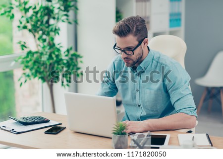 Profile side view photo of professional, concentrated, intelligent attractive bearded geek man in black spectacles sits in style, stylish denim blue shirt at the wooden table gazing into the monitor