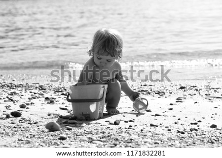 Small curious funny blonde child boy sitting on sea coast beach with wavy water sunny day outdoor playing with yellow plastic pail on natural background, horizontal picture