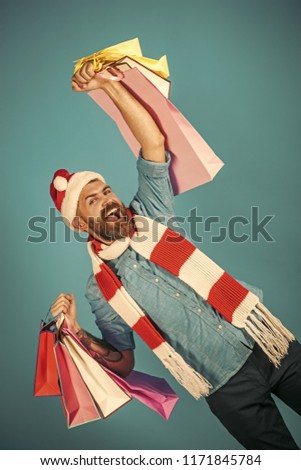 Christmas sale, black friday concept. Hipster hold shopping bags on blue background. New year, xmas presents. Man shopper smile in santa hat and scarf with winner gesture. Winter holidays celebration.