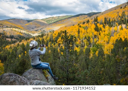 Woman sitting on a boulder taking a picture of the fall foliage with her phone