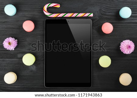 Office desk with macarons, lollipop, tablet-touch screen device and pink plants on black table background. Top view. Flat lay