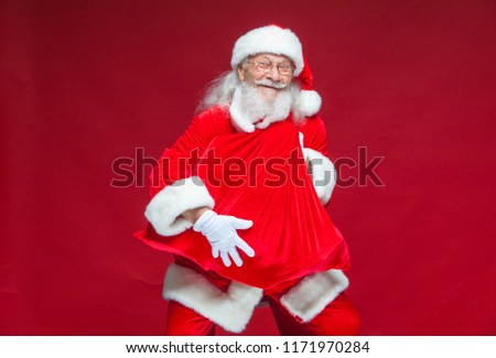 Christmas. A kind smiling Santa Claus is holding a red bag with presents in front of him embracing it. Isolated on red background.