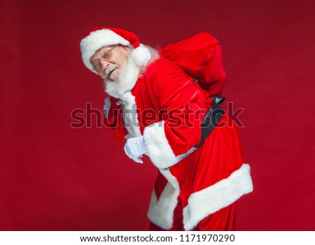 Christmas. Kind and tired Santa Claus in white gloves carries a red bag with gifts over his shoulder. Isolated on red background.