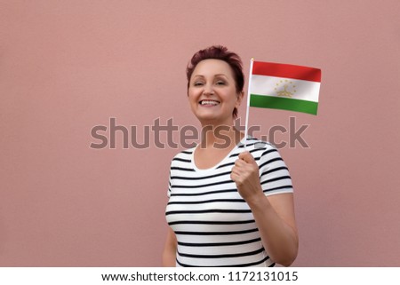 Tajikistan flag. Woman holding Tajikistan flag. Nice portrait of middle aged lady 40 50 years old with a national flag over pink wall background outdoors.