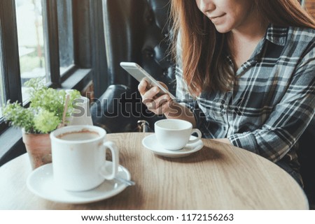 Closeup image of an asian woman holding , using and touching a smart phone with coffee cups on table in cafe