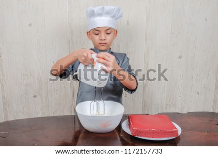 Happy boy making whipped cream in glass bowl and preparing a red velvet cake.
