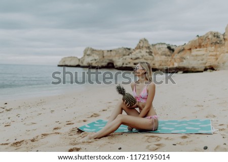 slim woman in bikini and sunglasses holding fresh pineapple near the sea