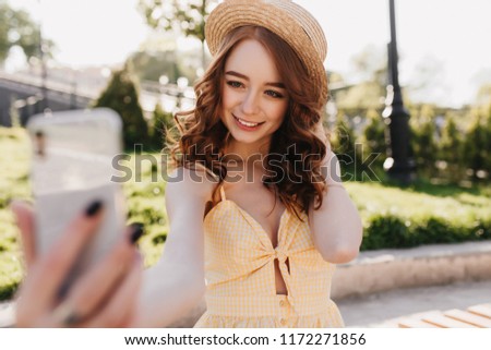 Debonair ginger lady in hat making selfie in green park. Outdoor photo of fascinating european woman with red hair taking picture of herself on nature background.