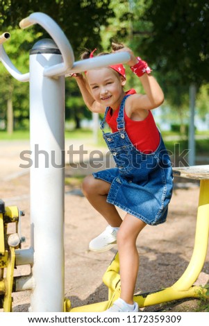 pretty little girl in denim suit and red bandana plays on children's playground. the kid is engaged on sports simulator outdoors in the park