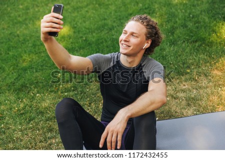 Smiling young sportsman listening to music with earphones while sitting on a fitness mat outdoors, holding mobile phone, taking a selfie