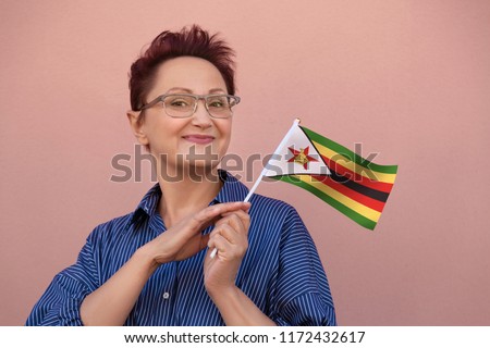 Zimbabwe flag. Woman holding Zimbabwe flag. Nice portrait of middle aged lady 40 50 years old with a national flag over pink wall on the street outdoors.