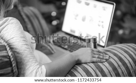 Closeup black and white photo of young woman sitting in armchair holding credit card and browsing online store on laptop