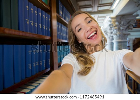 Excited young girl standing in traditional old library at bookshelves, smiling and laughing student making selfie on phone camera, having fun. Higher education and Student life concept.