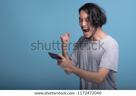 Excited young bearded man playing mobile games in blue background. Asian young hipster playing mobile game and win, half body shot. Young generation hipster relaxing concept. Royalty-Free Stock Photo #1172472040